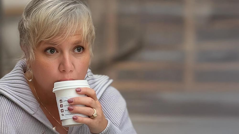 Woman with short blonde hair holding a coffee cup, looking off thoughtfully during a quiet outdoor moment.