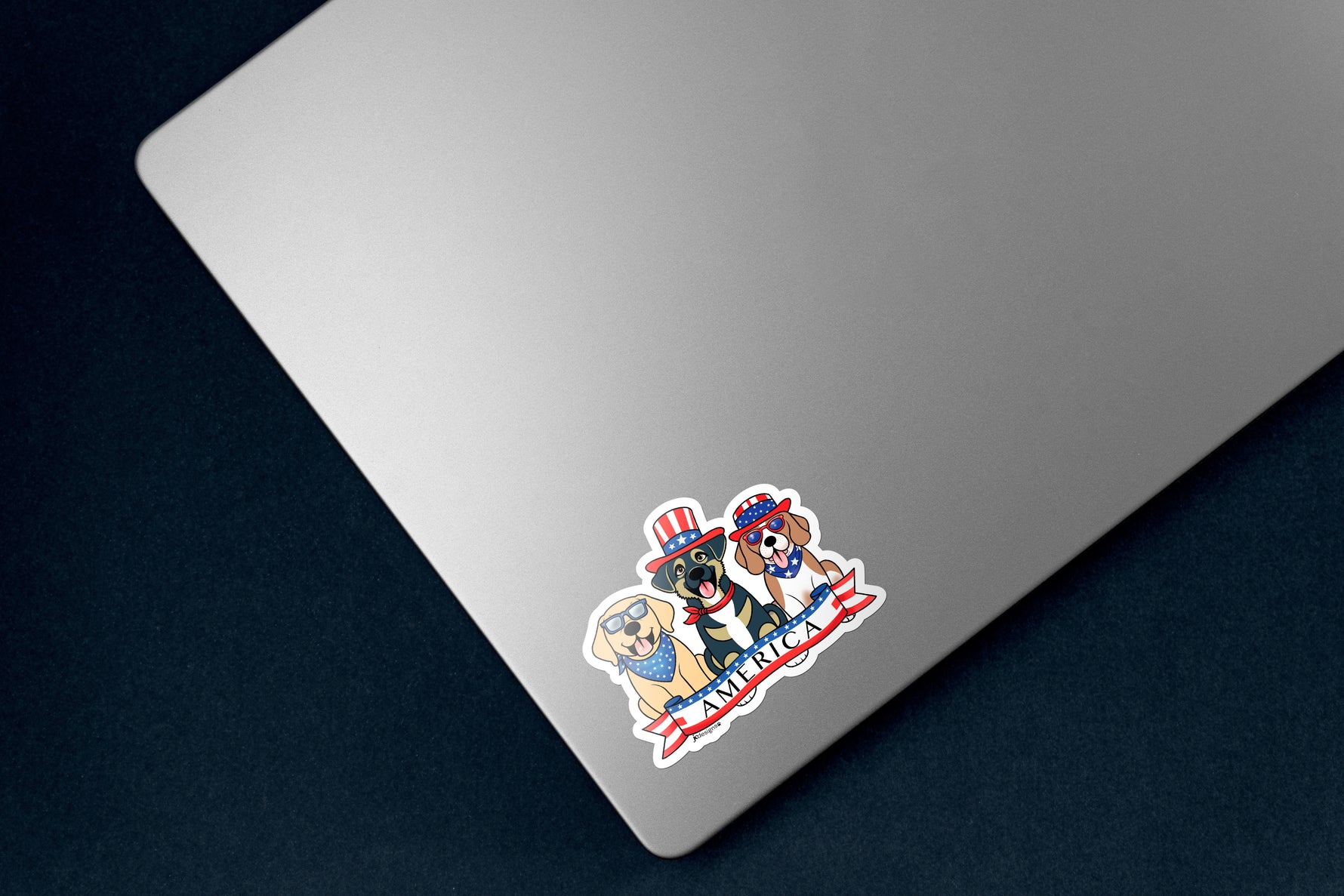 Sticker of three dogs - a Yellow Lab wearing a blue and white star bandana, a brown and tan mutt wearing a red, white, and blue USA flag top hat, and a brown and white Beagle wearing a red, white, and blue USA flag top hat and blue and white star bandana. A red, white, and blue banner with text 'America' is below the dogs. The sticker is on a silver laptop.