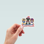 Hand holding a sticker of three dogs - a Yellow Lab wearing a blue and white star bandana, a brown and tan mutt wearing a red, white, and blue USA flag top hat, and a brown and white Beagle wearing a red, white, and blue USA flag top hat and blue and white star bandana. A red, white, and blue banner with text 'America' is below the dogs.