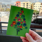 Hand holding card outdoors with colorful city buildings in background, the card is a Paw Print Christmas Tree card featuring blue orange pink green paws and yellow bones shaped like a Christmas Tree with a blue wavy line as the tree trunk on a green background