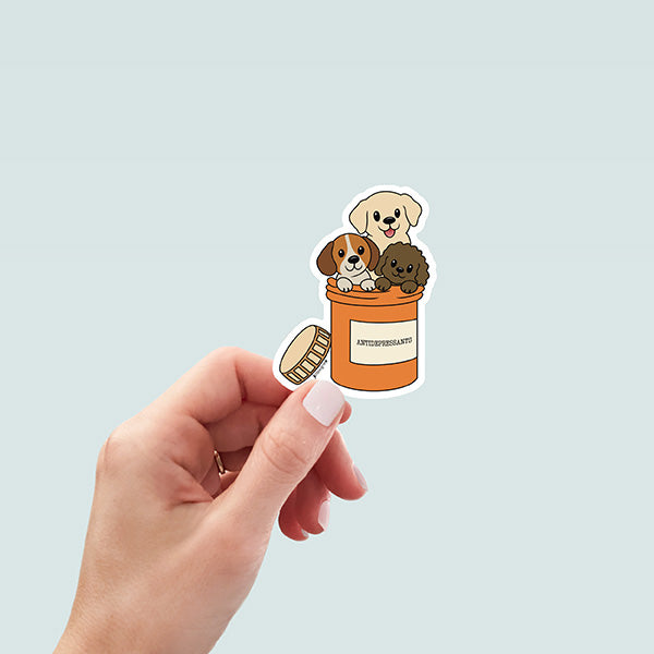hand holding cute sticker of three cartoon dogs - a brown curly labradoodle, yellow lab, and a red, tan, and white beagle - sitting inside an orange pill bottle with lid resting on side of bottle, labeled "Antidepressants"