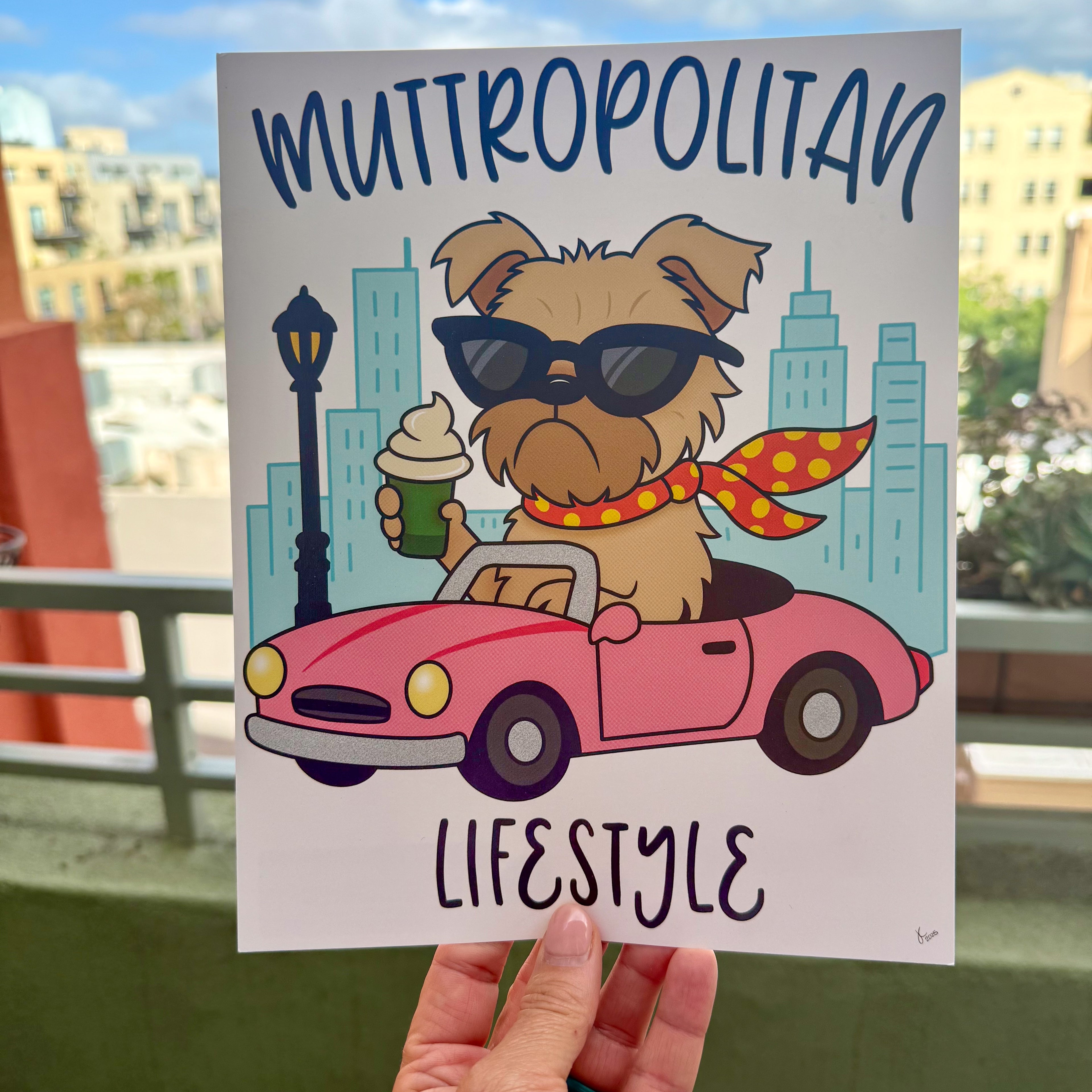 hand holding Muttropolitan Lifestyle art print outside with city buildings in the background, the art print is featuring a tan Brussels Griffon dog wearing black sunglasses and a red and yellow polk-a-dot scarf blowing in the wind, holding a puppucino in a green coffee cup and driving a pink convertible car with city high rise buildings and black light post in the background, the words muttropolitan are rounded on top the image and lifestyle is on the bottom, overall image has a white background