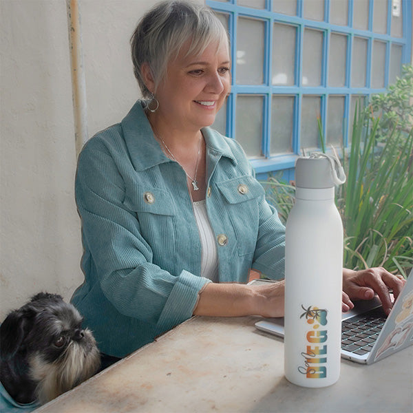 Jillann sitting at table on her laptop, a water bottle with San Diego dog paw sticker on it sits next to the computer and Oz is sitting next to Jillann looking up at the computer