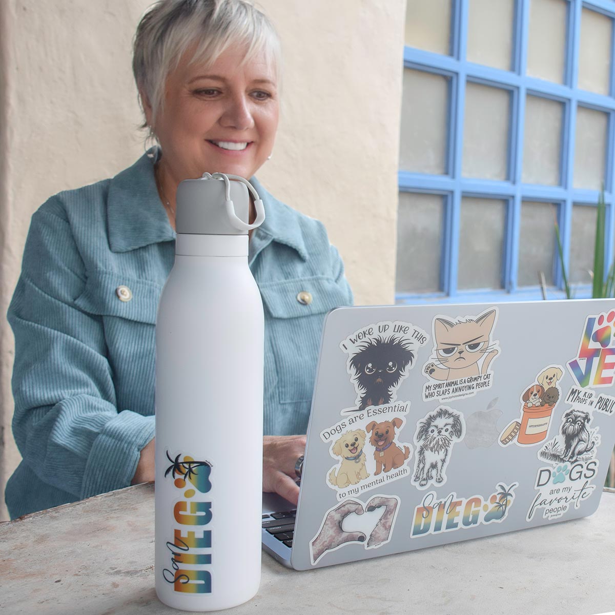 Jillann smiling while working on laptop decorated with cute cat and dog stickers, white water bottle with dog sticker sits next to computer, stickers by JC Pet Designs