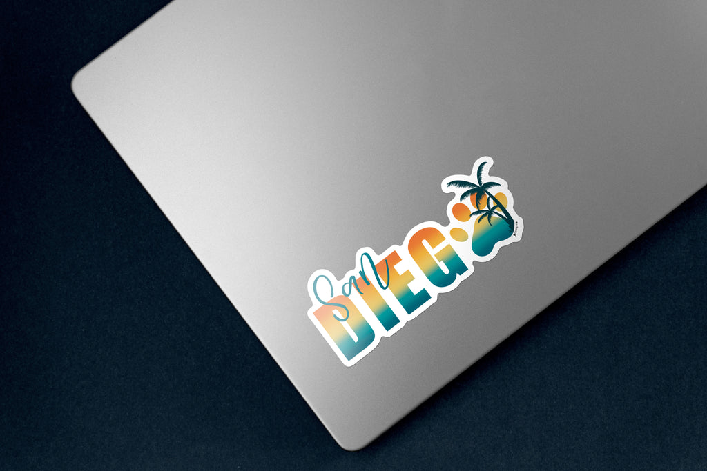 Sticker design with the words 'San Diego' in a bold gradient font transitioning from teal to yellow to orange, resembling a sunset. The word 'San' is written in a teal script font above the 'D' in 'Diego'. The 'O' is replaced with a large paw print in matching gradient colors, overlaid with a silhouette of a palm tree. The design is outlined in white. The sticker is on a silver laptop.