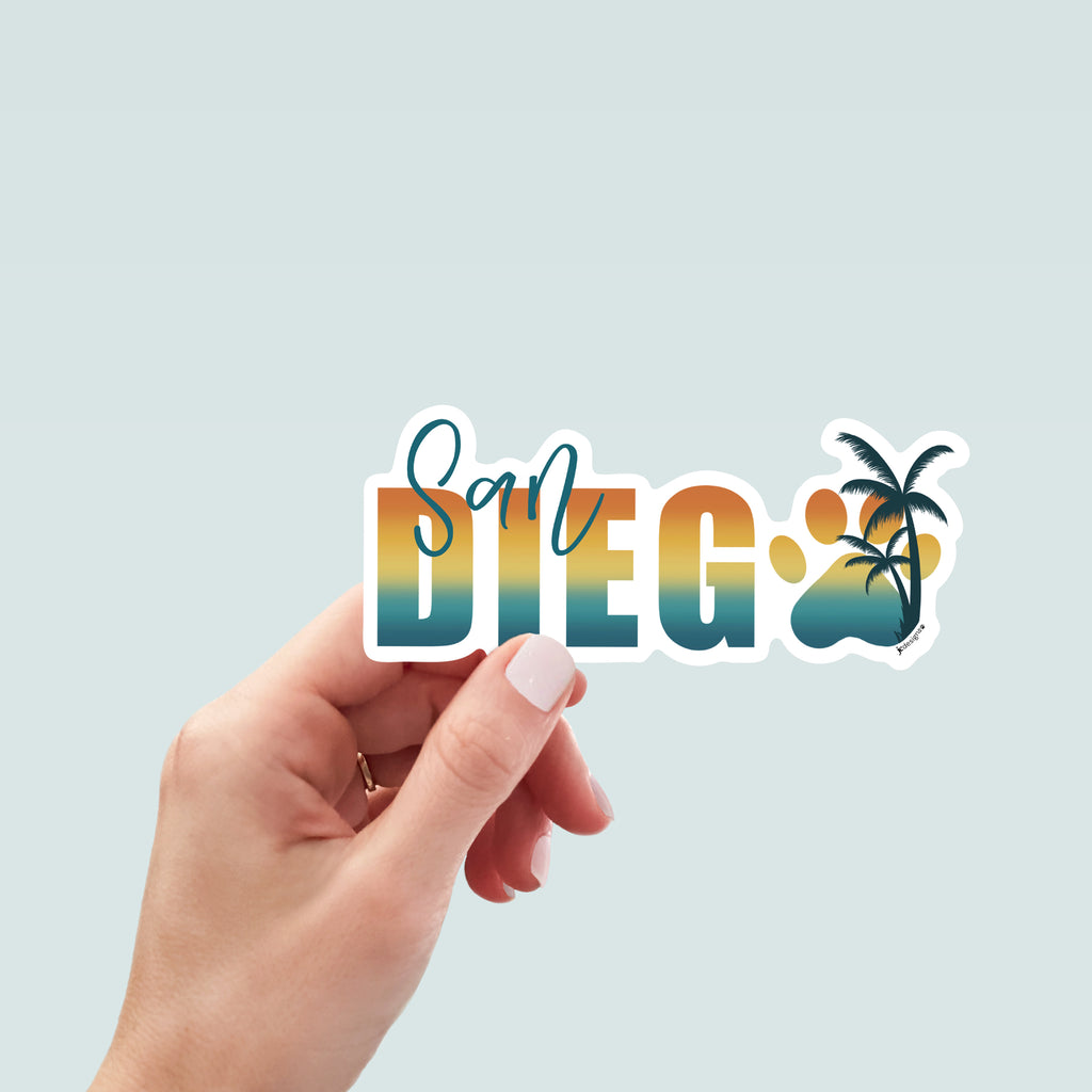 Hand holding a sticker design with the words 'San Diego' in a bold gradient font transitioning from teal to yellow to orange, resembling a sunset. The word 'San' is written in a teal script font above the 'D' in 'Diego'. The 'O' is replaced with a large paw print in matching gradient colors, overlaid with a silhouette of a palm tree. The design is outlined in white.