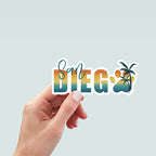 Hand holding a sticker design with the words 'San Diego' in a bold gradient font transitioning from teal to yellow to orange, resembling a sunset. The word 'San' is written in a teal script font above the 'D' in 'Diego'. The 'O' is replaced with a large paw print in matching gradient colors, overlaid with a silhouette of a palm tree. The design is outlined in white.