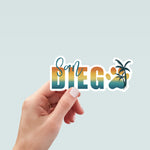 Hand holding a sticker design with the words 'San Diego' in a bold gradient font transitioning from teal to yellow to orange, resembling a sunset. The word 'San' is written in a teal script font above the 'D' in 'Diego'. The 'O' is replaced with a large paw print in matching gradient colors, overlaid with a silhouette of a palm tree. The design is outlined in white.