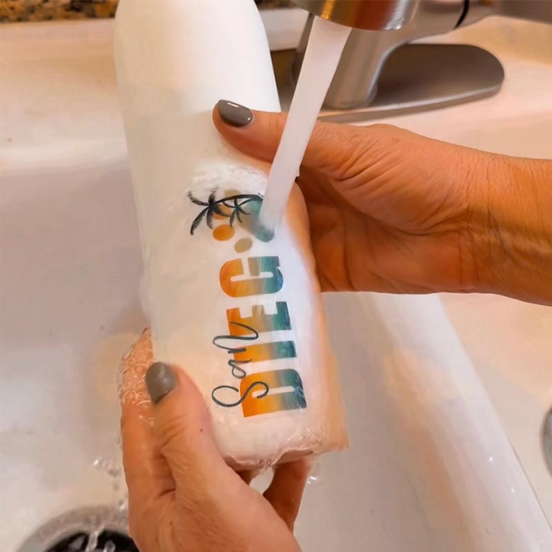Waterproof pet-themed vinyl sticker on a white bottle being rinsed under a running faucet, demonstrating durability and long-lasting quality from JC Pet Designs