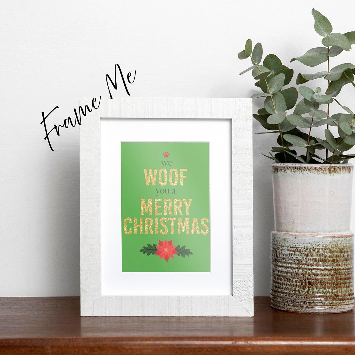 Framed We Woof You a Merry Christmas holiday card featuring Woof and Merry Christmas in gold-glitter-style text, we and you a are in a dark green text, a red paw print is on top of the card and a red poinsettia is on the bottom of the card, the background is green, frame not included in purchase