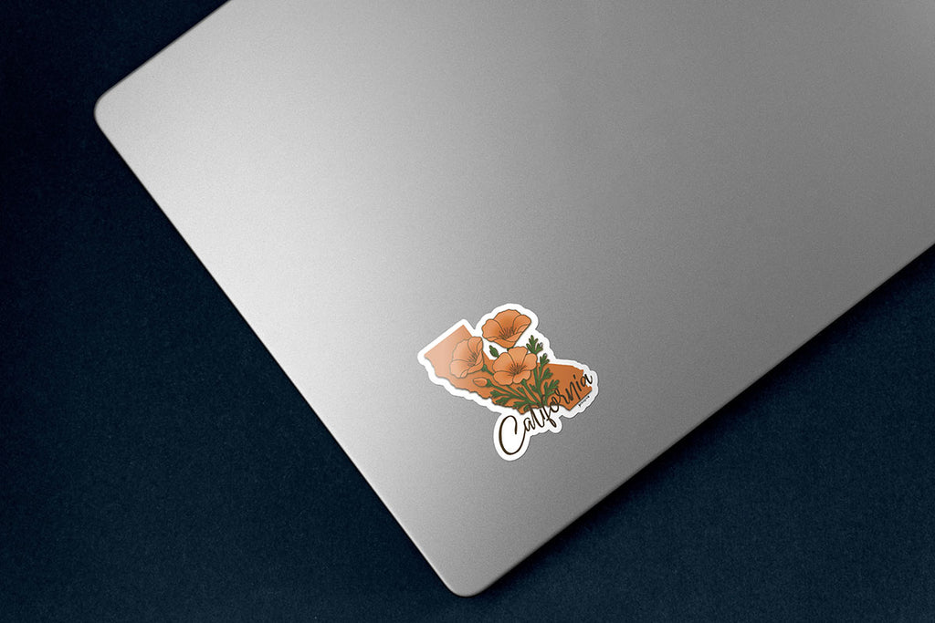 California state-shaped sticker featuring vibrant orange poppy flowers extending out of state outline with elegant script text reading "California" at bottom of sticker, white outline die cut, sticker is on lower left corner of silver laptop