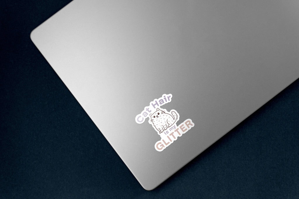 Sticker of a fluffy white cartoon cat covered in scattered strands of cat hair. Above the cat, the phrase 'Cat Hair' is written in soft purple text, with 'is my' is smaller matching text below the cat. The word 'GLITTER' is prominently displayed at the bottom in large, sparkling textured letters. The design is outlined in white. the 3 inch sticker is on a silver laptop. 