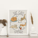 framed The Cats' Timetable art print featuring four illustrated cats lounging, stretching, napping, and playing around a clock design, text The Cat's is arched over the top and text Timeable arched under the bottom, the image is on a light tan background, frame does not come with purchase