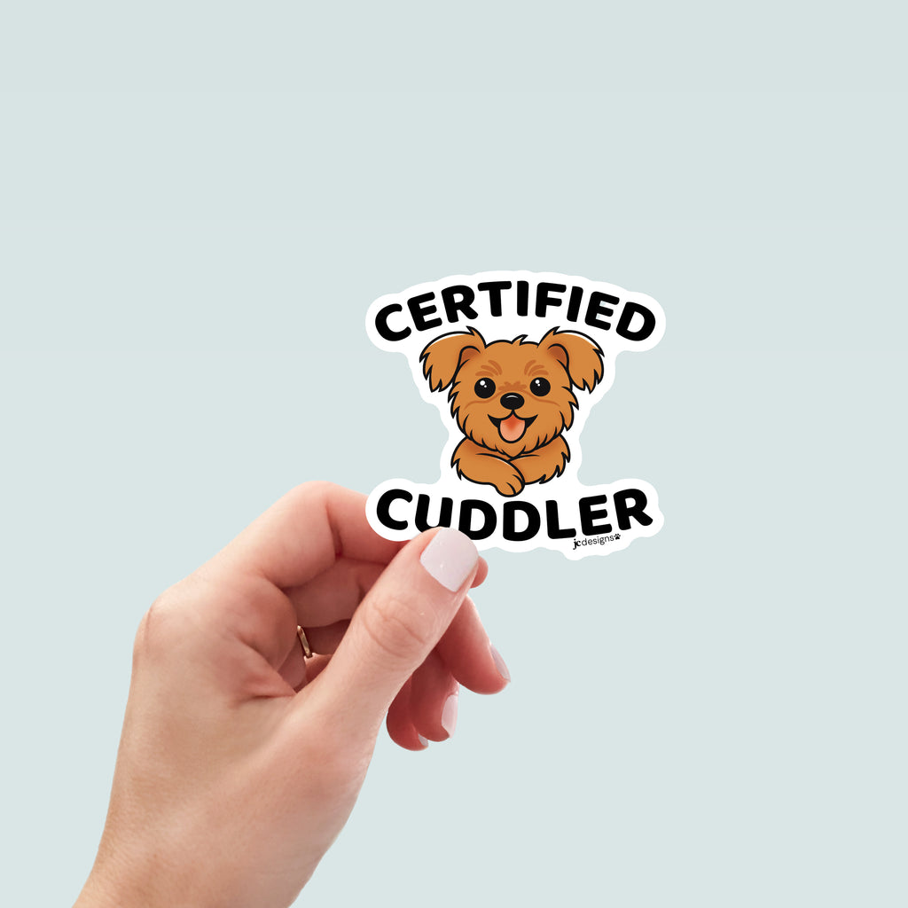 Hand holding a sticker design featuring a cute, fluffy brown dog with a happy expression, tongue out, and front paws crossed. The text 'Certified' arched above the dog in bold black letters, and 'Cuddler' is written below in matching font. The design is outlined in white.