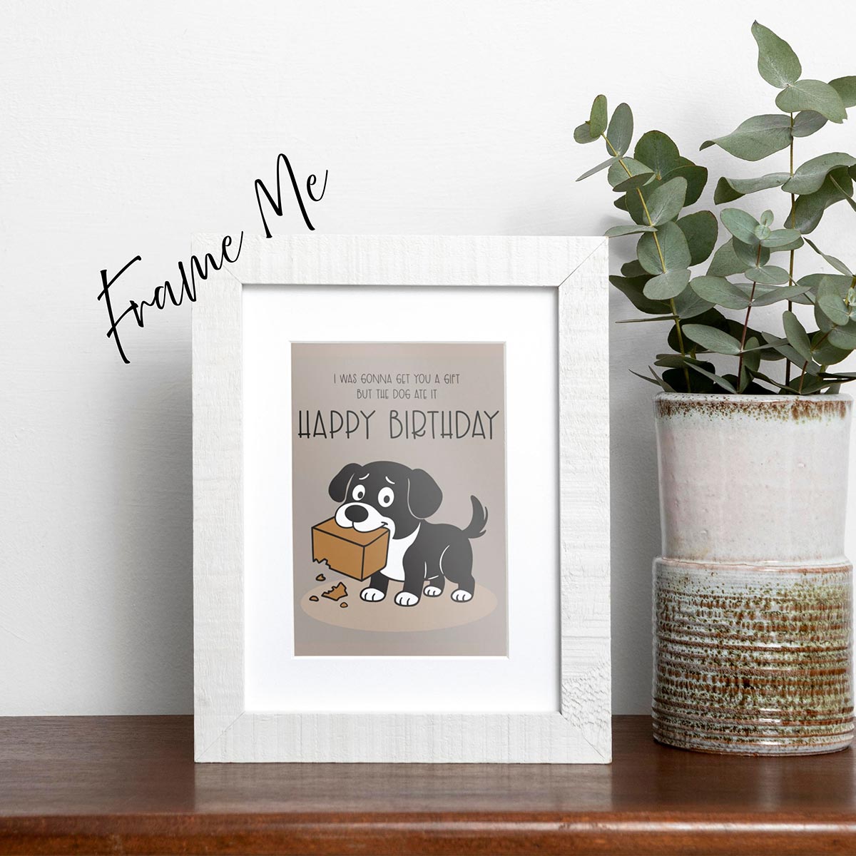 framed The Dog Ate It Birthday card featuring a cartoon black and white dog holding a chewed-up gift box with the text that reads, "I was gonna get you a gift but the dog ate it, Happy Birthday", the card is on a brown background, frame not included in purchase