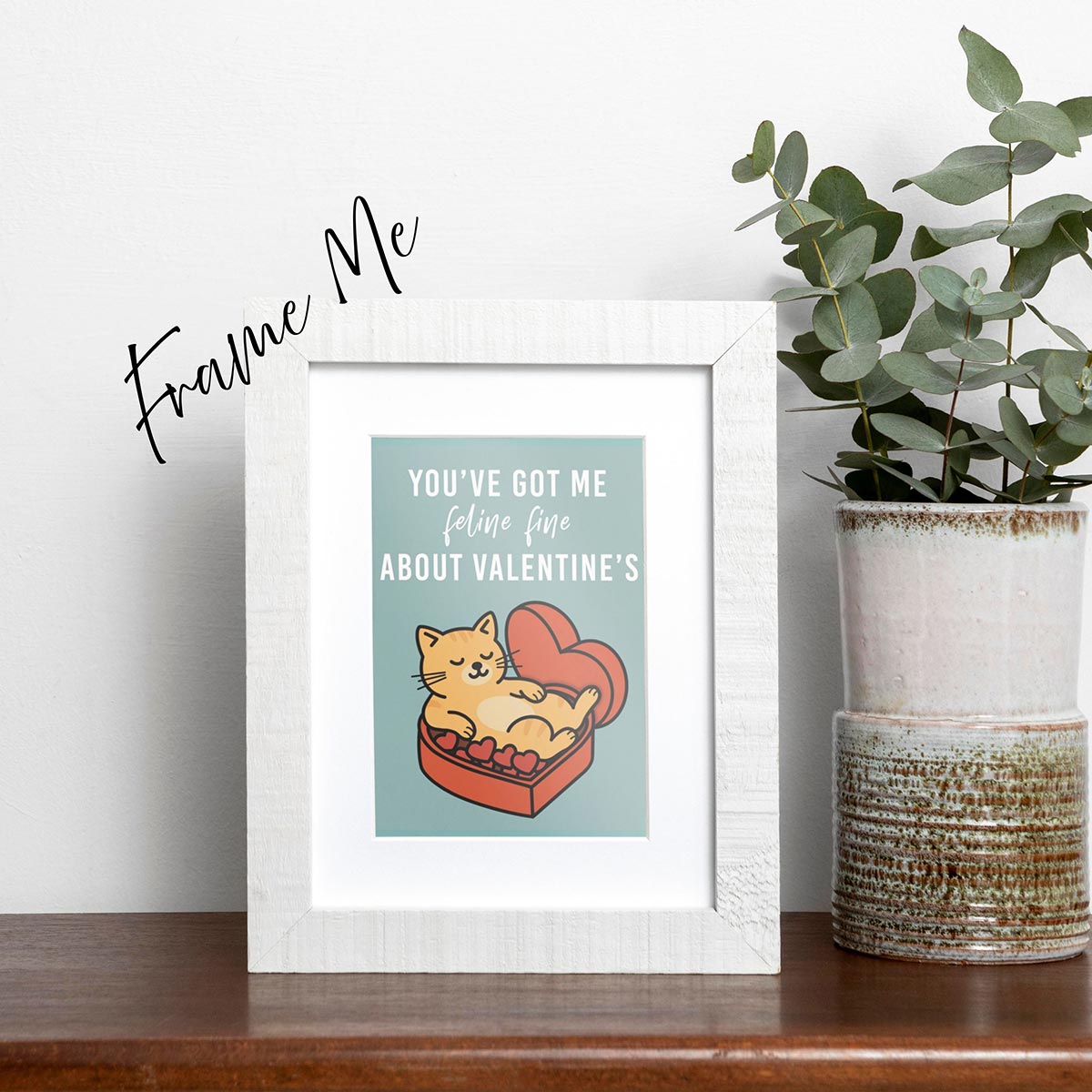 framed Feline Fine Valentine's Day card features an orange cat relaxing on a box of red heart chocolates inside a red heart-shaped chocolate box, text in white on the top of the card reads "You've got me Feline Fine about Valentine's", the card has a muted teal background