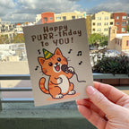 hand holding card outdoors with colorful city buildings in the background, the Happy Purr-thday birthday card features an orange cat with tan belly standing up with microphone in left paw and green & yellow party hat on and musical notes floating around cat, text on top of card reads "Happy Purr-thday to You", the card has a muted light brown background