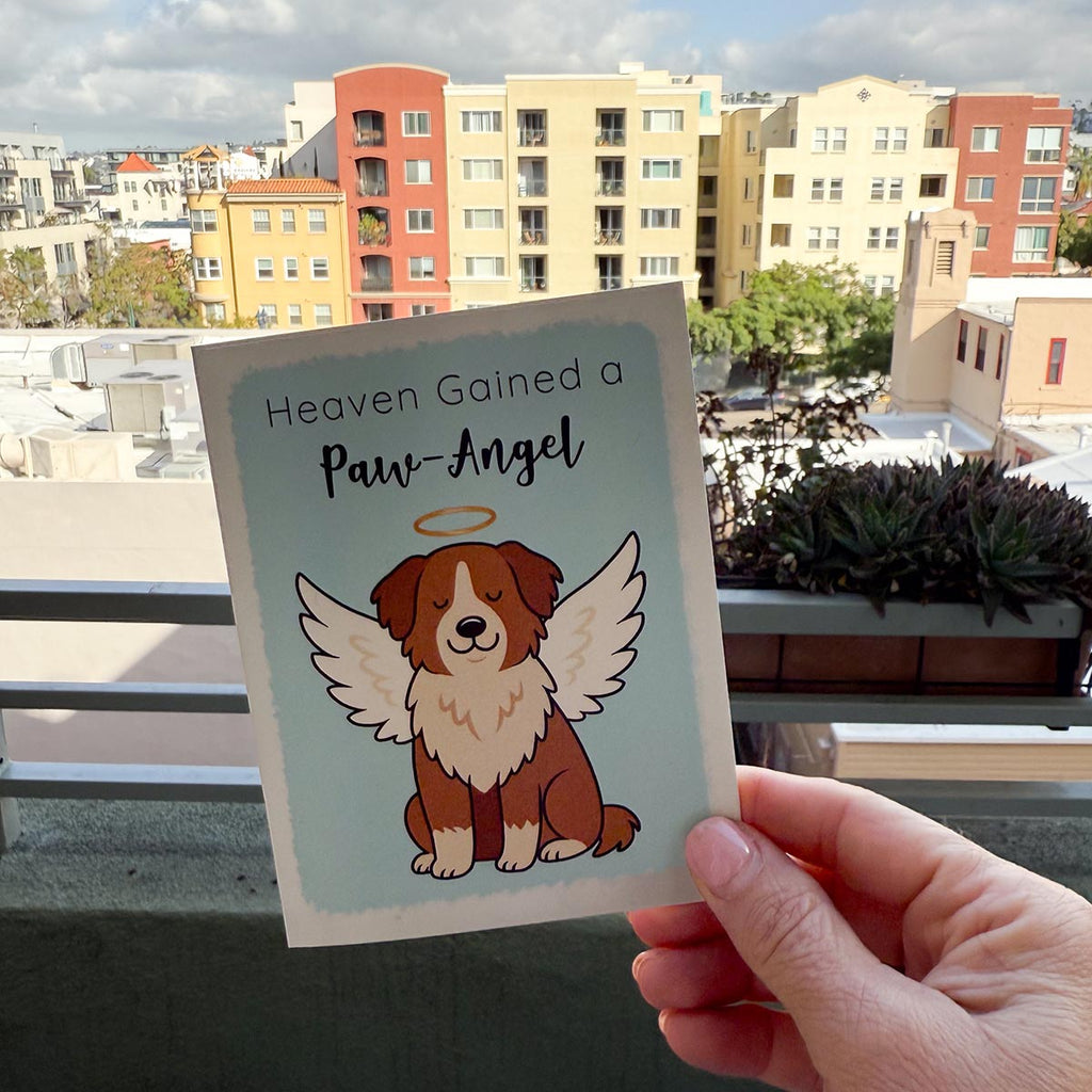 hand holding card outdoors with colorful city buildings in background, the Heaven Gained a Paw-Angel pet sympathy card features a brown and white dog sitting up facing forward with angel wings and a golden halo on a soft blue background, text on top of card reads "Heaven Gained a Paw-Angel"