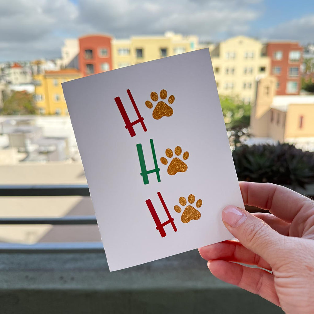 hand holding card outdoors with colorful city buildings in background, the card is the Ho Ho Ho Christmas Holiday card featuring red and green letters with gold paw prints in place of the "O" of all 3 Ho Ho Ho's, on a white background