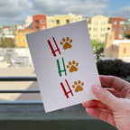 hand holding card outdoors with colorful city buildings in background, the card is the Ho Ho Ho Christmas Holiday card featuring red and green letters with gold paw prints in place of the "O" of all 3 Ho Ho Ho's, on a white background