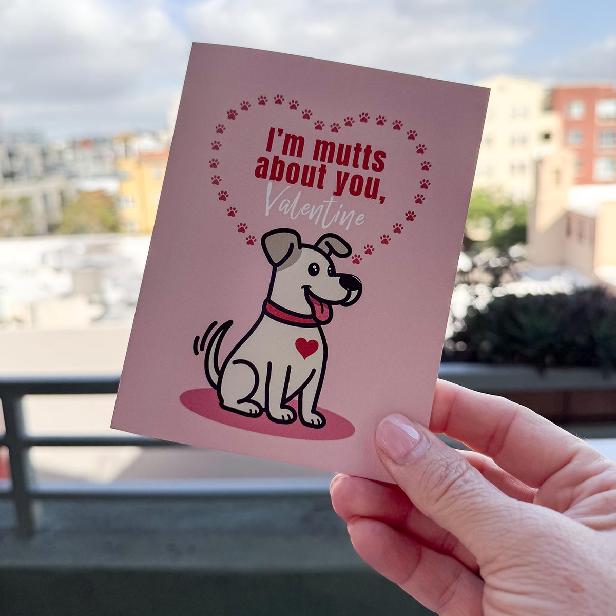 hand holding card outdoors with colorful city buildings in background, the Tuna Time card features a cute orange cat sitting on a can of tuna, text on top of card reads "You Make Me Purr Louder than Tuna Time", the card has a muted pink background