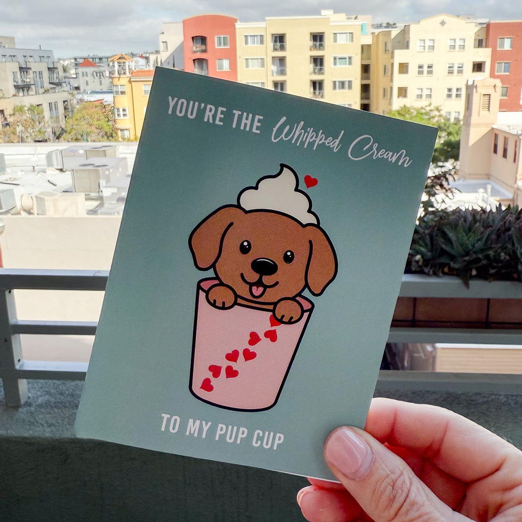 hand holding card outdoors with colorful city buildings in the background, the Pup Cup Valentine's Day card features a cute brown puppy stinking tongue out with whipped cream on its head and peaking out of a pink cup decorated with red hearts, a small red heart  is floating above whipped cream, text at top of card reads "You're the Whipped Cream" and the text at the bottom of the card reads "to my Pup Cup", the card has a muted teal color
