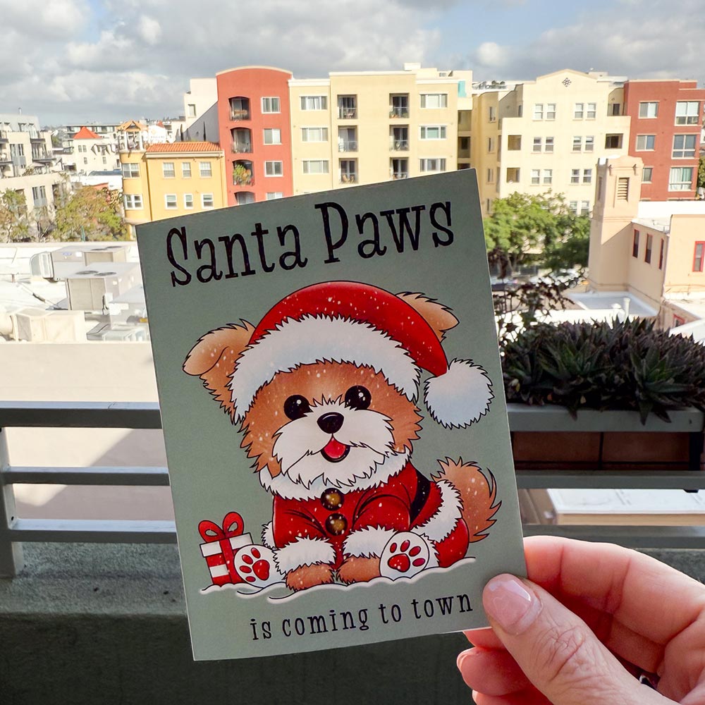 hand holding card outdoors with colorful city buildings in background, the Santa Paws Christmas card features an adorable fluffy brown puppy in a Santa suit and hat sitting in snow next to a red and white wrapped gift, puppy's paw pads are red, text on top of card reads "Santa Paws", text on bottom of card reads "is coming to town", card has a muted teal background