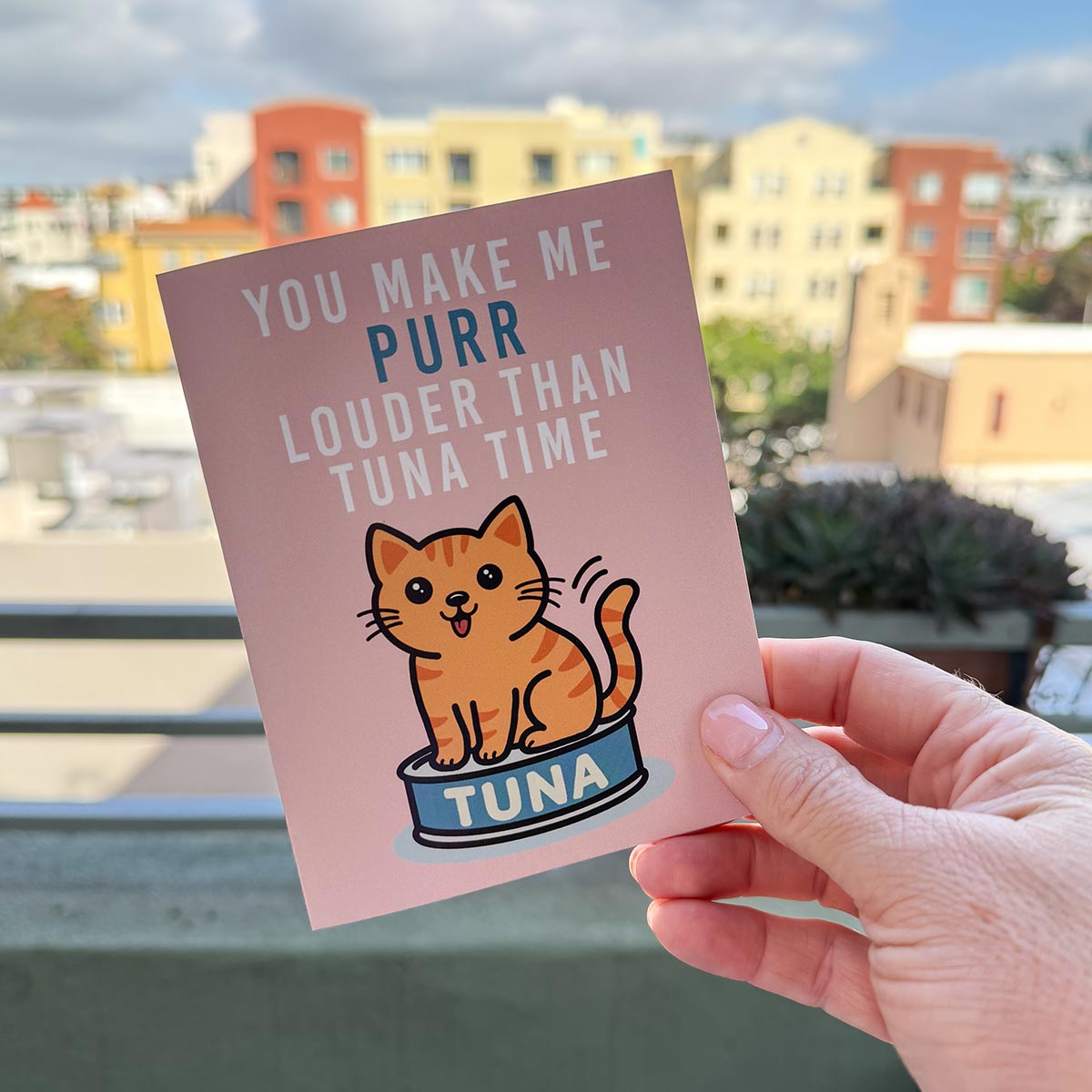 hand holding card outdoors with colorful city buildings in the background, the Tuna Time card features a cute orange cat sitting on a can of tuna, text on top of card reads "You Make Me Purr Louder than Tuna Time", the card has a muted pink background