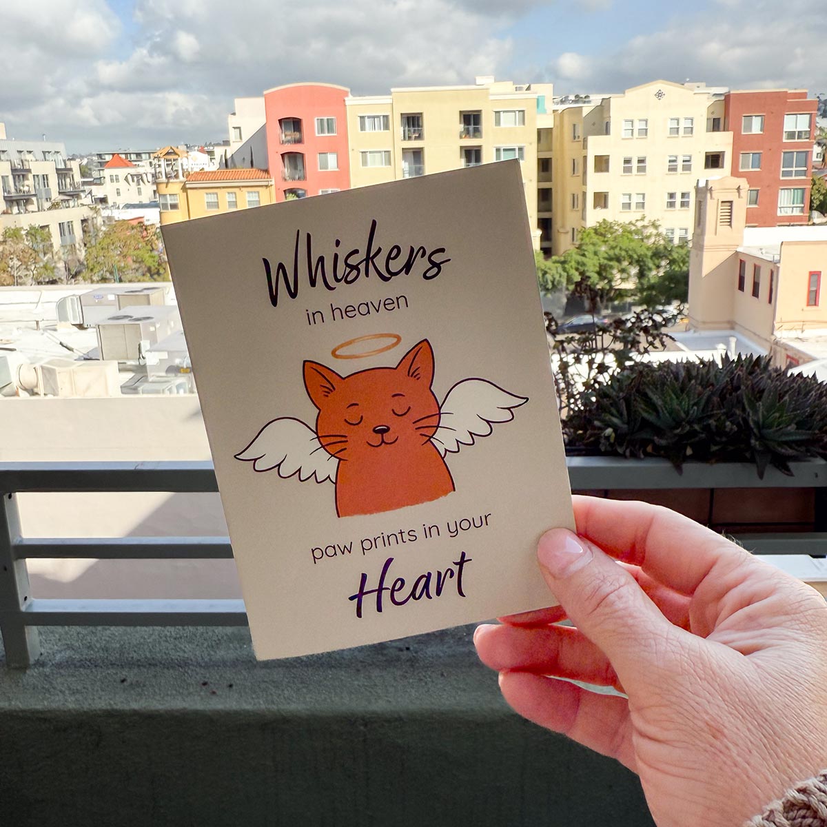 hand holding card outdoors with colorful city buildings in background, the Whiskers in Heaven pet loss sympathy card features an orange cat with angel wings and gold halo, text reads Whiskers in heaven at top of card and paw prints in your heart at bottom of card, card has a muted light brown background