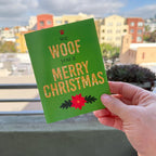 hand holding card outdoors with colorful city buildings in the background, the card is the We Woof You a Merry Christmas holiday card featuring Woof and Merry Christmas in gold-glitter-style text, we and you a are in a dark green text, a red paw print is on top of the card and a red poinsettia is on the bottom of the card, the background is green