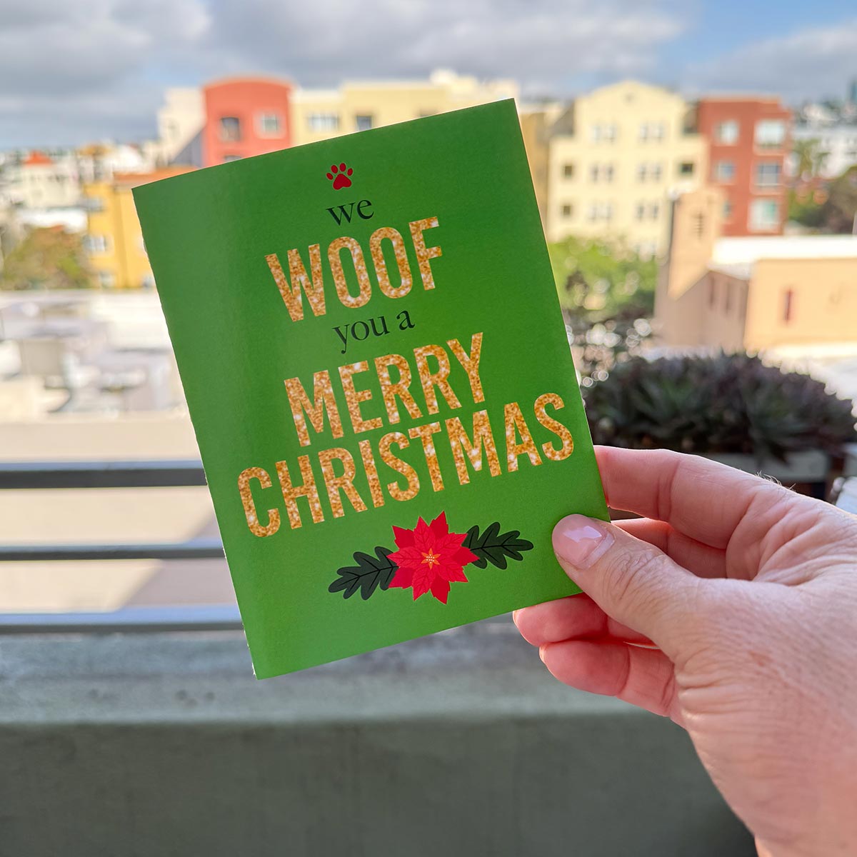 hand holding card outdoors with colorful city buildings in the background, the card is the We Woof You a Merry Christmas holiday card featuring Woof and Merry Christmas in gold-glitter-style text, we and you a are in a dark green text, a red paw print is on top of the card and a red poinsettia is on the bottom of the card, the background is green