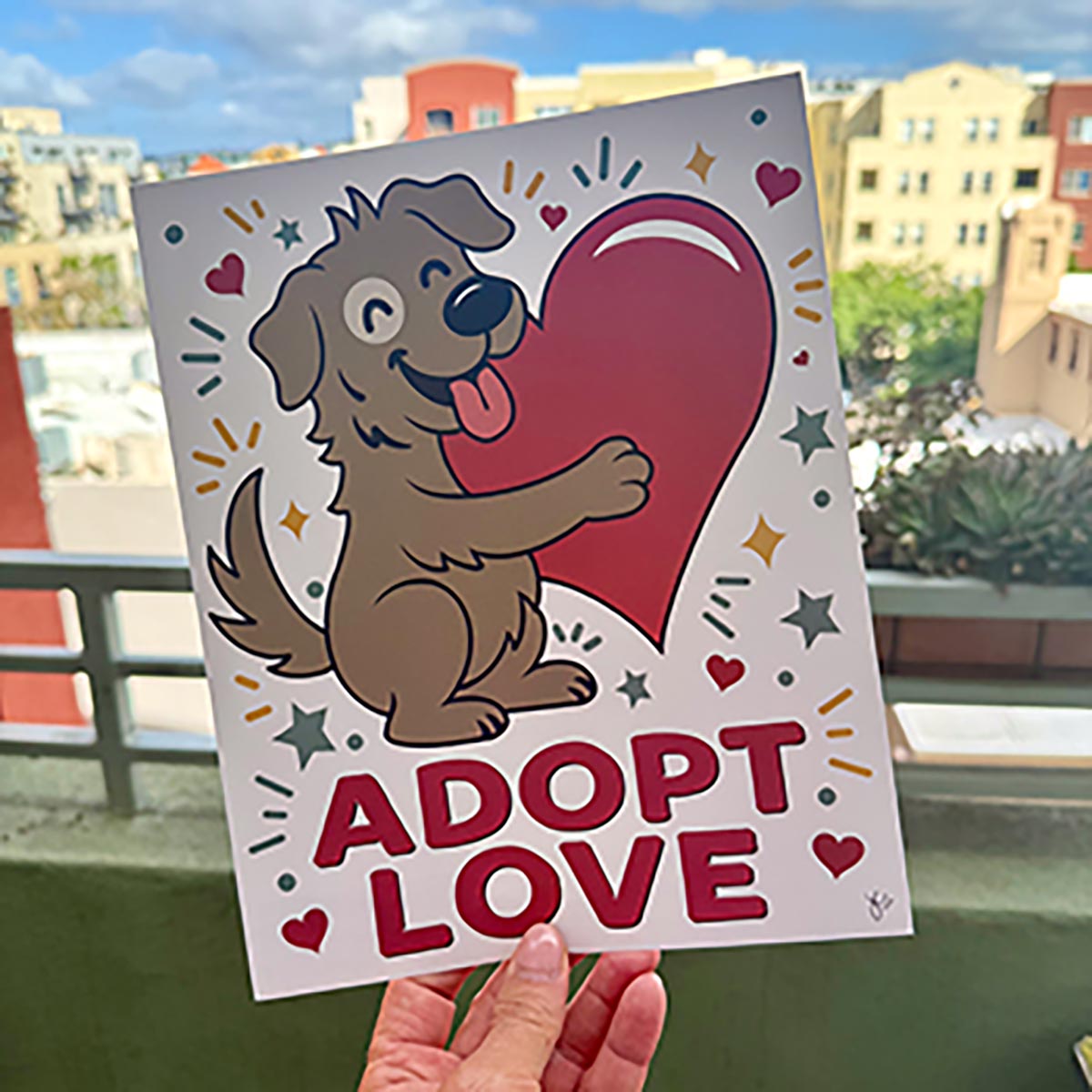 Hand holding Adopt Love art print outdoors with colorful city buildings in background, Adopt Love art print is with smiling brown dog with white patch around right eye hugging a red heart surrounded by yellow diamonds, teal stars, red hearts, teal dots, and teal & yellow lines on a white background