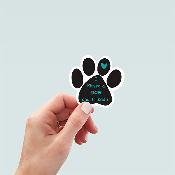 hand holding black paw-shaped sticker with text in the main paw pad in teal reading "I kissed a Dog and I liked it" and teal heart on second paw pad from right