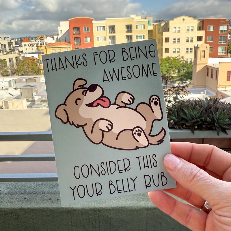 hand holding card outdoors with colorful city buildings in the background, the Thanks for Being Awesome greeting card features a happy brown dog with a light tan belly lying on its back with its tongue sticking out, text on the card reads "Thanks for Being Awesome, Consider This Your Belly Rub", card has a muted teal background