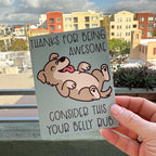 hand holding card outdoors with colorful city buildings in the background, the Thanks for Being Awesome greeting card features a happy brown dog with a light tan belly lying on its back with its tongue sticking out, text on the card reads "Thanks for Being Awesome, Consider This Your Belly Rub", card has a muted teal background