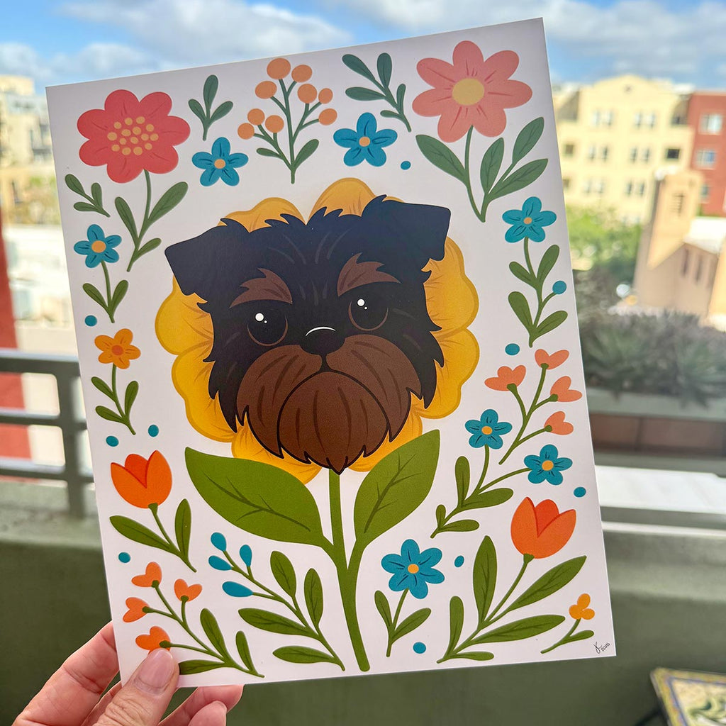 hand holding art print outdoors with colorful city buildings in the background, the Brussels Griffon flower art print features a black and tan Griffon dog's face framed by a yellow flower surrounded by orange and teal flowers and green leaves and stems on a white background