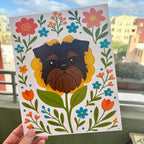 hand holding art print outdoors with colorful city buildings in the background, the Brussels Griffon flower art print features a black and tan Griffon dog's face framed by a yellow flower surrounded by orange and teal flowers and green leaves and stems on a white background