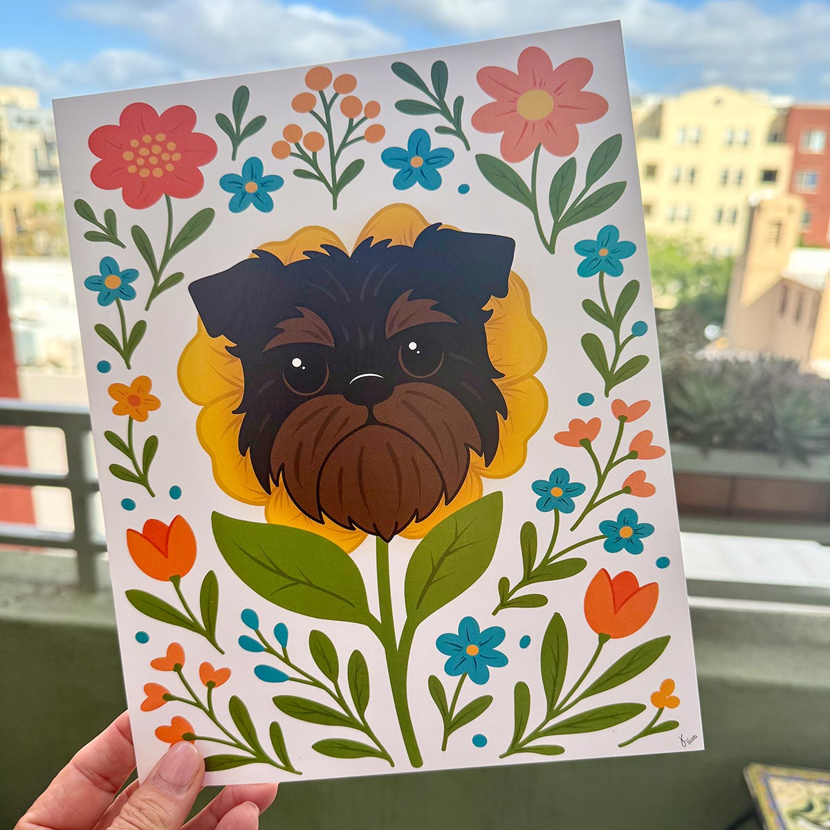 hand holding art print outdoors with colorful city buildings in the background, the Brussels Griffon flower art print features a black and tan Griffon dog's face framed by a yellow flower surrounded by orange and teal flowers and green leaves and stems on a white background