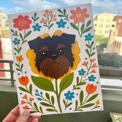 hand holding art print outdoors with colorful city buildings in the background, the Brussels Griffon flower art print features a black and tan Griffon dog's face framed by a yellow flower surrounded by orange and teal flowers and green leaves and stems on a white background