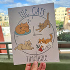 hand holding art print outdoors with city buildings in the background, the The Cats' Timetable art print features four illustrated cats lounging, stretching, napping, and playing around a clock design, text The Cat's is arched over the top and text Timeable arched under the bottom, the image is on a light tan background