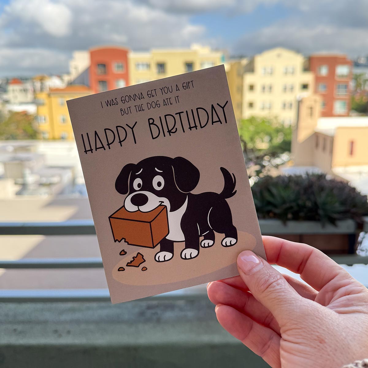 hand holding card outdoors with colorful city buildings in background, the The Dog Ate It Birthday card features a cartoon black and white dog holding a chewed-up gift box with the text that reads, "I was gonna get you a gift but the dog ate it, Happy Birthday", the card is on a brown background