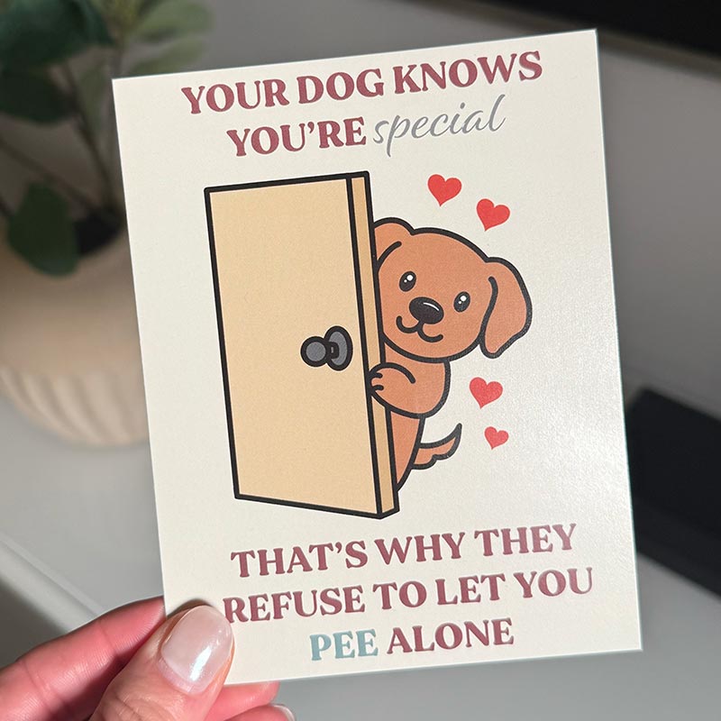 hand holding Illustrated greeting card showing a smiling dog peeking out from behind a door with hearts, reading “Your dog knows you’re special — that’s why they refuse to let you pee alone”
