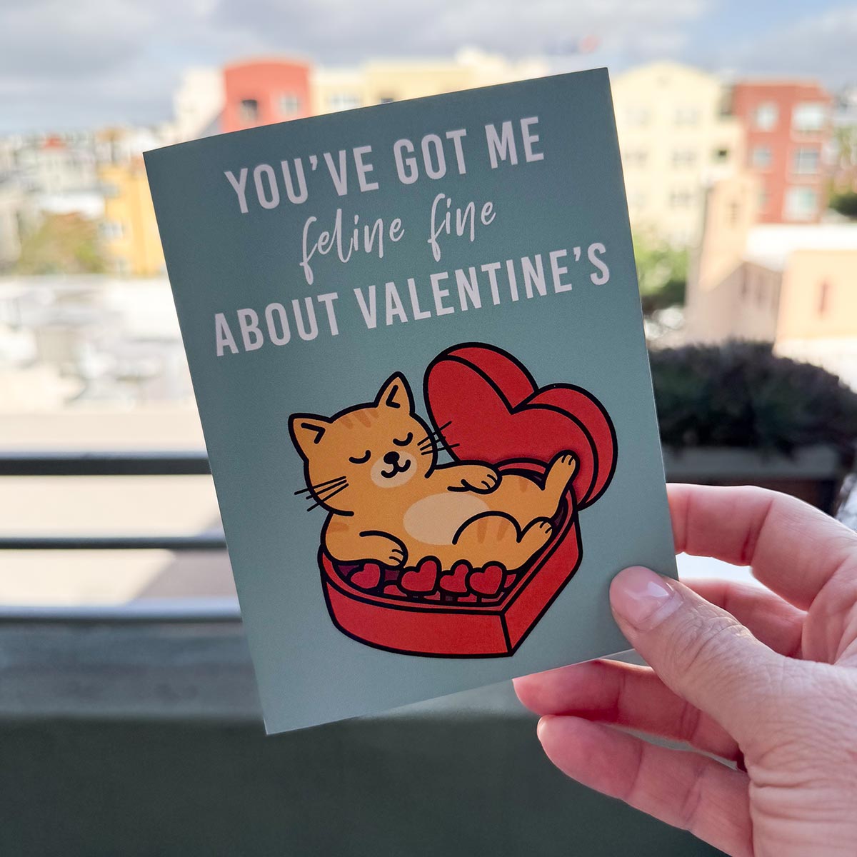 hand holding card outdoors with colorful city buildings in background, the Feline Fine Valentine's Day card features an orange cat relaxing on a box of red heart chocolates inside a red heart-shaped chocolate box, text in white on the top of the card reads "You've got me Feline Fine about Valentine's", the card has a muted teal background