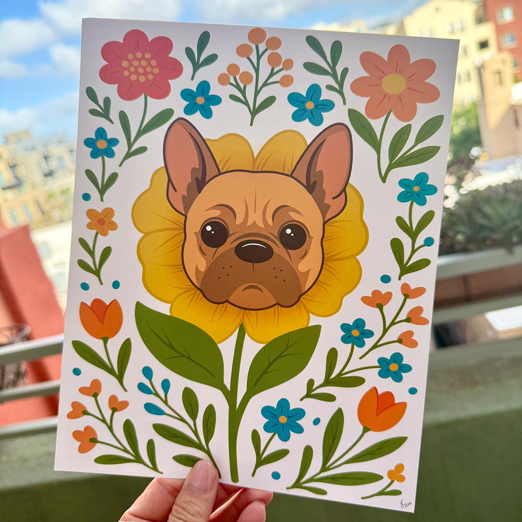 hand holding art print outdoors with city buildings in the background, French Bulldog flower art print features a brown and tan frenchie face framed by a yellow flower surrounded by orange and teal floral blooms and green leaf stems, print is on a white background