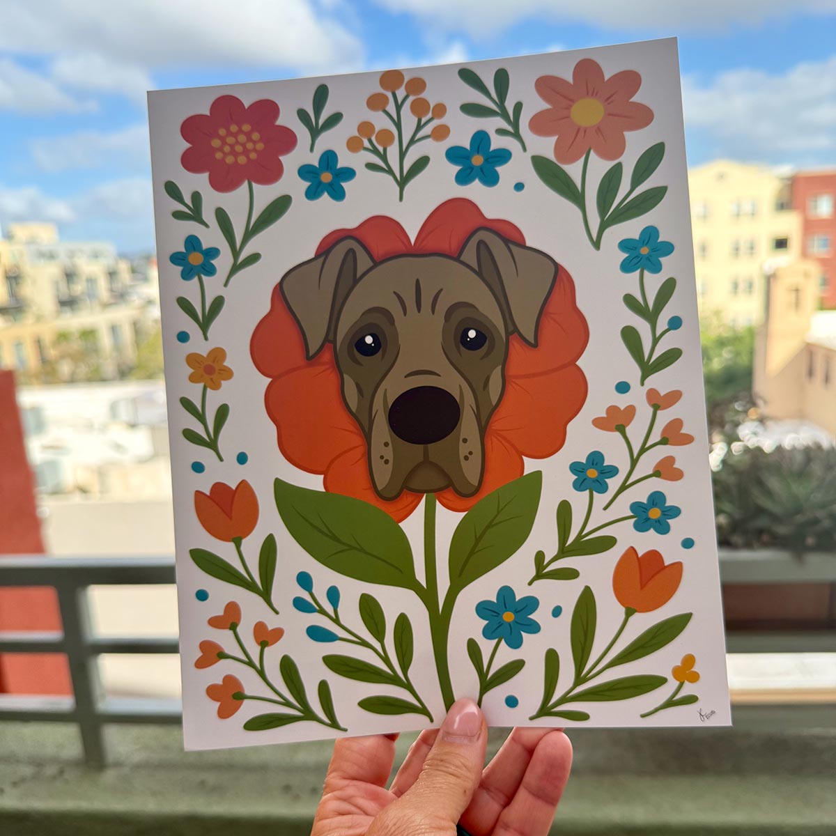 hand holding art print outdoors with colorful city buildings in the background, the Great Dane flower art print features a gray Great Dane's face framed by an orange flower surrounded by orange and teal flowers and green stems and leaves, on a white background