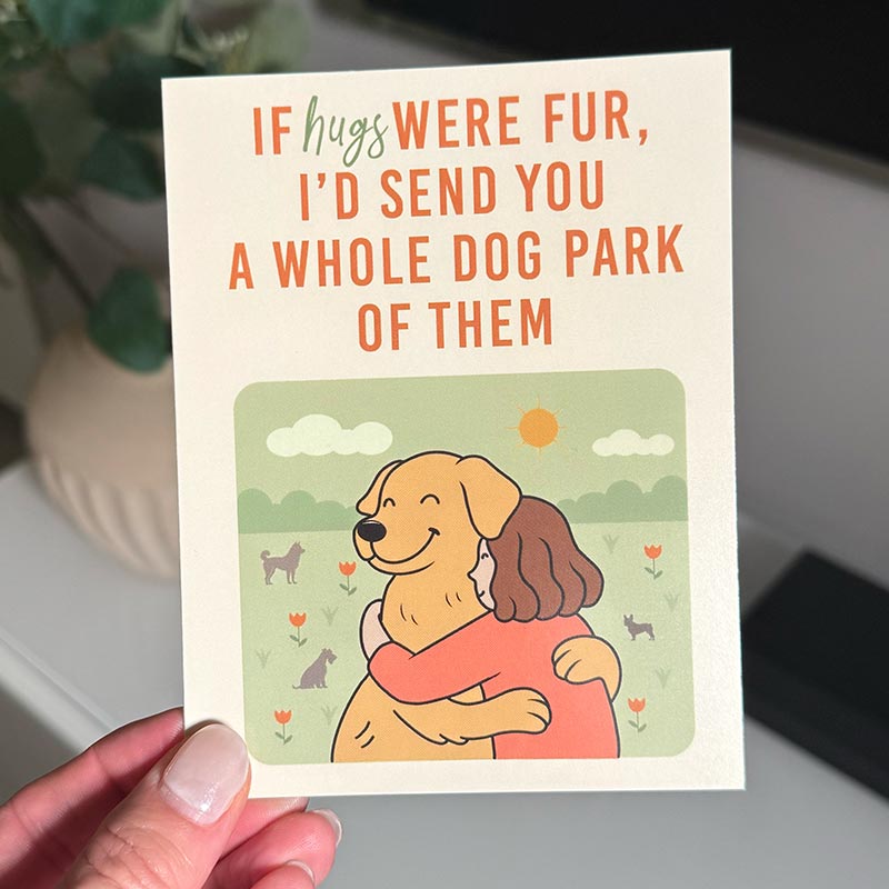 hand holding Illustrated greeting card showing a person hugging a dog in a park with the text “If hugs were fur, I’d send you a whole dog park of them”