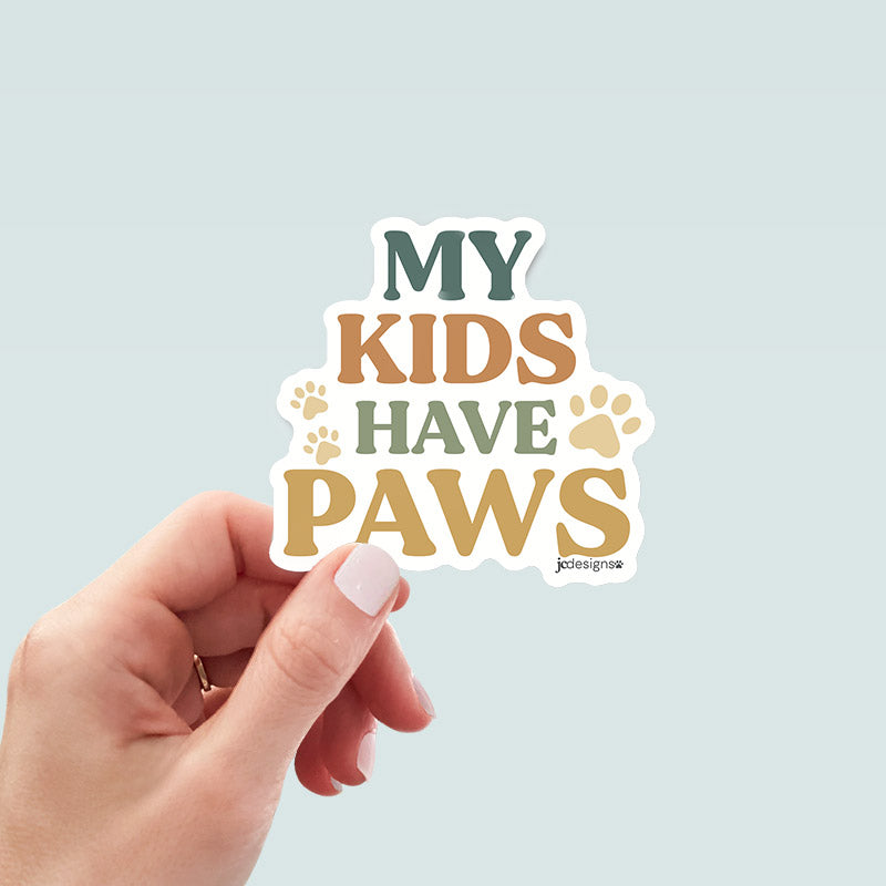 hand holding my kids have paws sticker with bold hand-lettered text and paw print details for proud pet parents, cat dads, dog moms