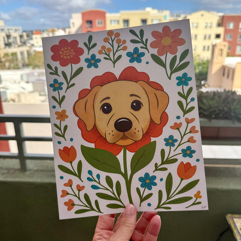 hand holding art print outdoors with colorful city buildings in the background, the Labrador flower art print features a yellow lab dog's face surrounded by orange teal and yellow flowers and green stems leaves on a white background