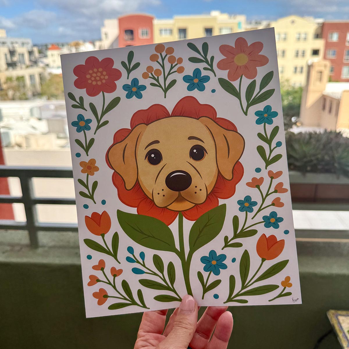 hand holding art print outdoors with colorful city buildings in the background, the Labrador flower art print features a yellow lab dog's face surrounded by orange teal and yellow flowers and green stems leaves on a white background