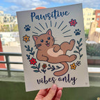 Person holding art print outdoors with city buildings in the background, the Pawsitive Vibes Only art print features a happy cartoon brown cat surrounded in a circle of red and yellow flowers and black paw prints with words Pawsitive on top and vibes on bottom print is on a light muted green background